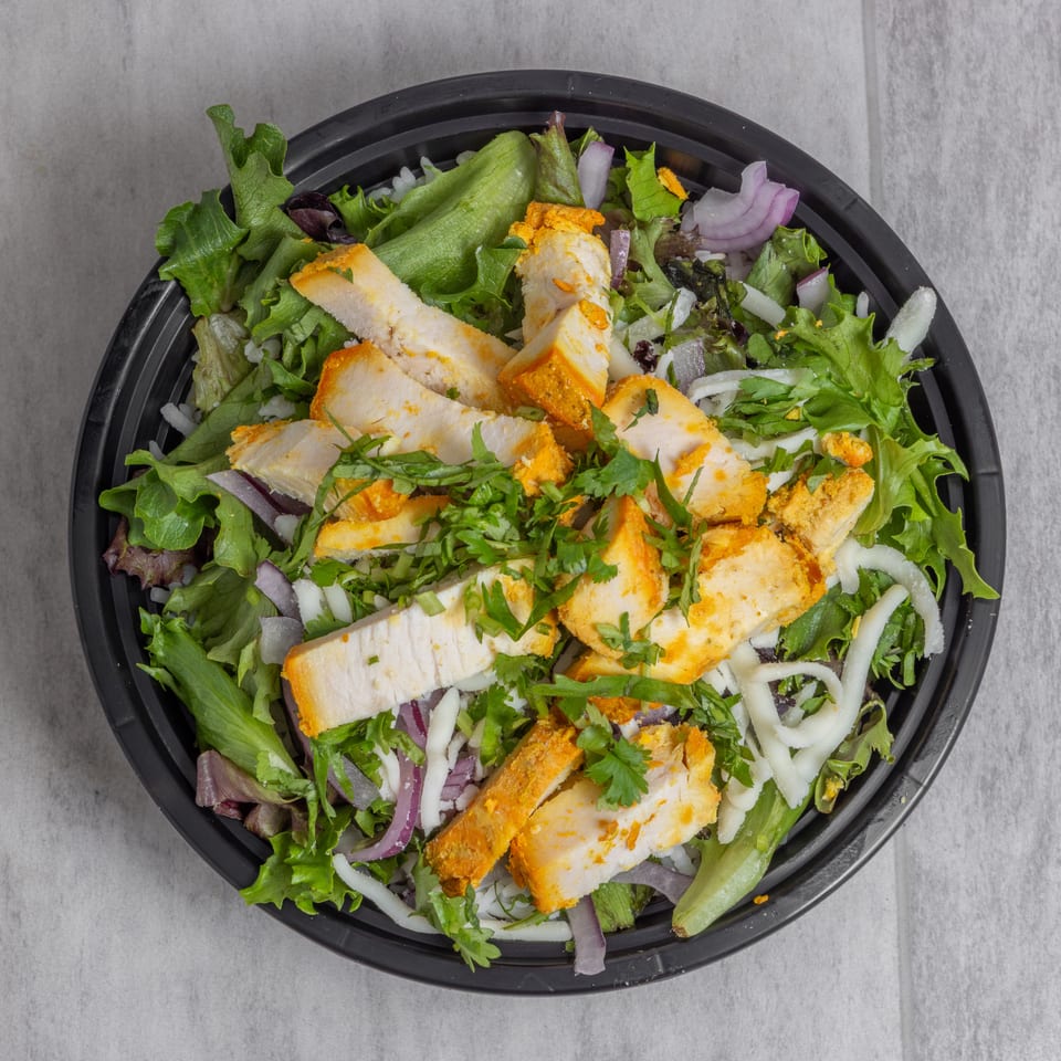Overhead view of a mixed green salad topped with sliced grilled chicken, red onion, shredded cheese, and fresh herbs.