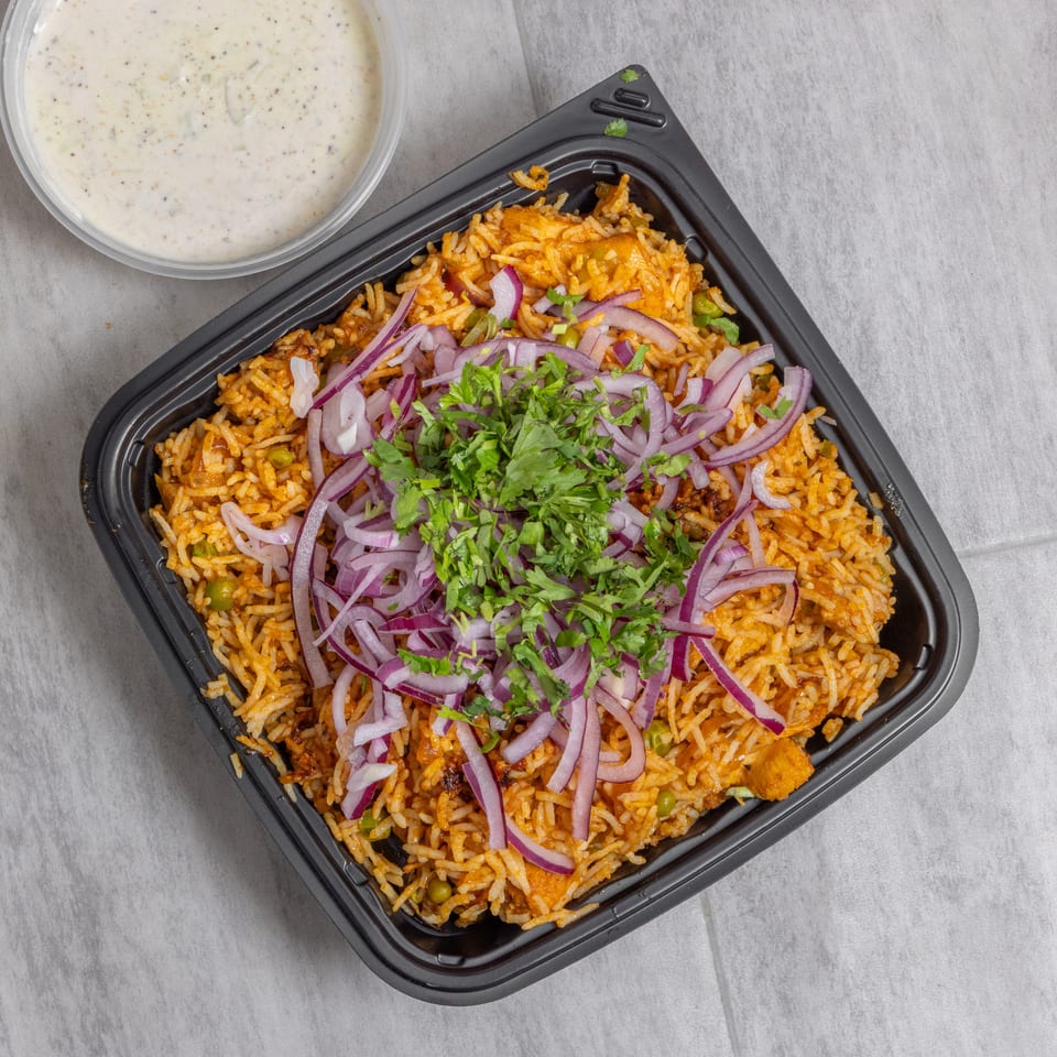 Spiced rice with chicken, peas, red onions, and cilantro in a black takeaway container, served with creamy yogurt sauce.