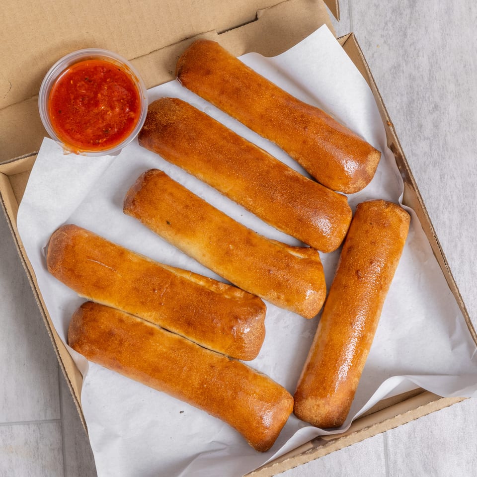 Open cardboard box holding golden-brown fried rolls with a cup of red dipping sauce.